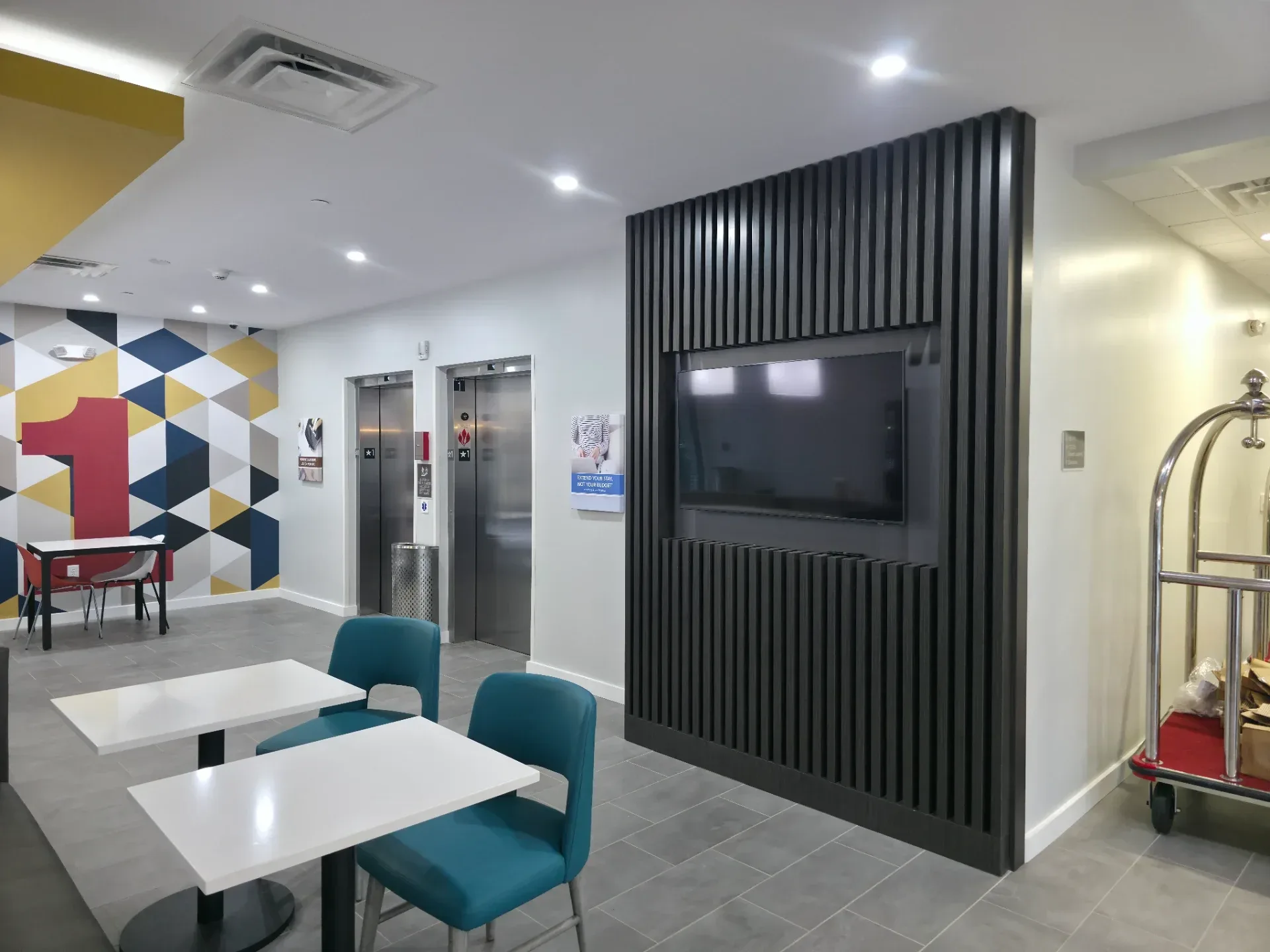 Dark slatted TV feature wall in hotel lobby adjacent to elevator bank, with white dining tables and teal chairs in view
